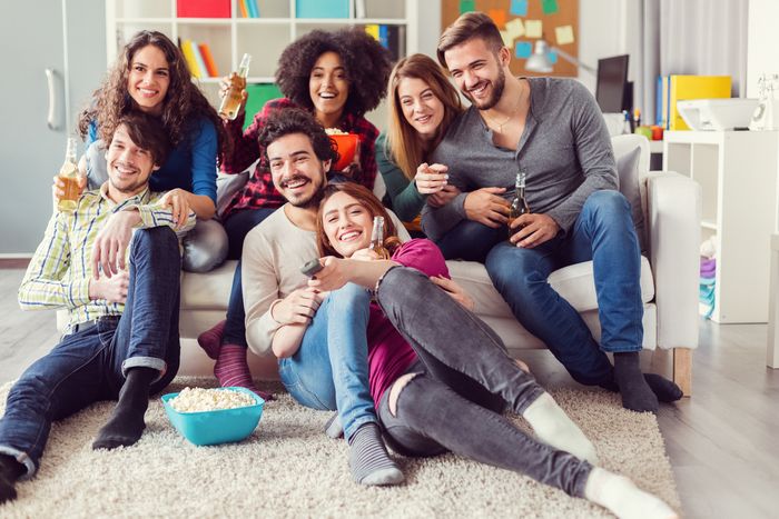 A group of seven people in their young twenties crowd around a couch and on the floor, drinking beer, laughing, and eating popcorn. A young woman sitting on the carpet in the front holds a TV remote.